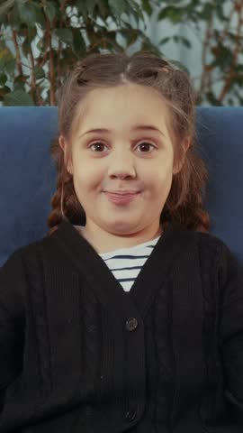 A girl sits on a blue chair, smiling playfully. The girl has braided hair and wears a black sweater over a striped shirt, with green foliage in the background.