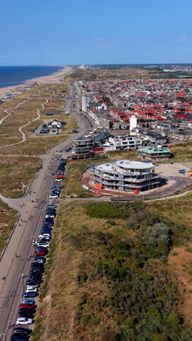 Vertical aerial drone video shot looking over the coastal boulevard and seaside resort architecture of Katwijk aan Zee, South Holland, Netherlands. The camera movement establishes a high-angle perspective tracing a long line of parked cars along the dune road and boulevard. In the foreground, a modern circular building complex under active construction sits surrounded by coastal sand dunes covered in grass and green vegetation. Beyond the road, red-tiled residential roofs, coastal apartment blocks, and historic town architecture spread out toward the horizon. On the left, sprawling dune walking paths lead toward the North Sea beach shoreline under a clear blue sky on a bright summer afternoon. High-resolution stock media highlighting Dutch coastal real estate development, urban construction projects, beach resort infrastructure, and dune protection landscapes. Ideal for vertical social media reels, mobile video production, travel stories, commercial advertising, and broadcast media.