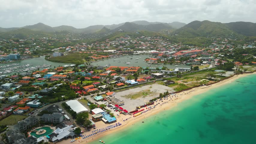 Wide elevated view over Reduit Beach in Gros Islet, Saint Lucia, with beach umbrellas, yachts moored in Rodney Bay Marina, orange roofed town and Monier mountain ridge behind