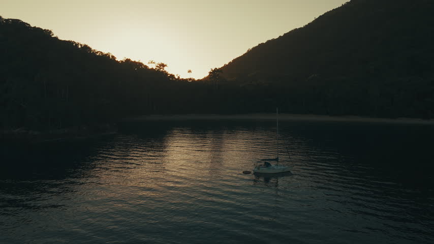 Aerial view of the sailing boat anchored in the calm lagoon with sun rising and shining from the mountains
