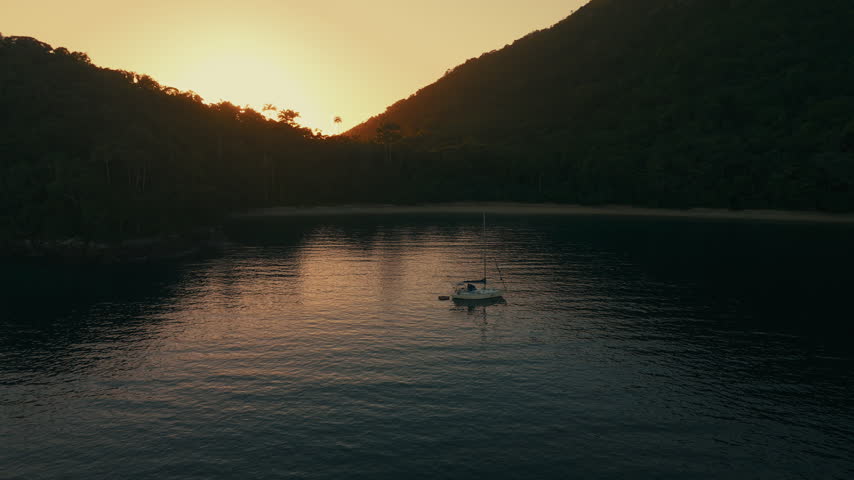 Aerial view of the sailing boat anchored in the calm lagoon with sun rising and shining from the mountains