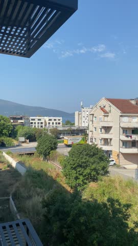 Elevated panning view of a Mediterranean coastal town with residential buildings and green hills under a blue sky. The camera pans slowly from left to right, showcasing apartment blocks, parked vehicles, and local streets. Forested mountains rise in the background beneath a clear sky. The scene captures the peaceful, sunny atmosphere of Tivat, Montenegro