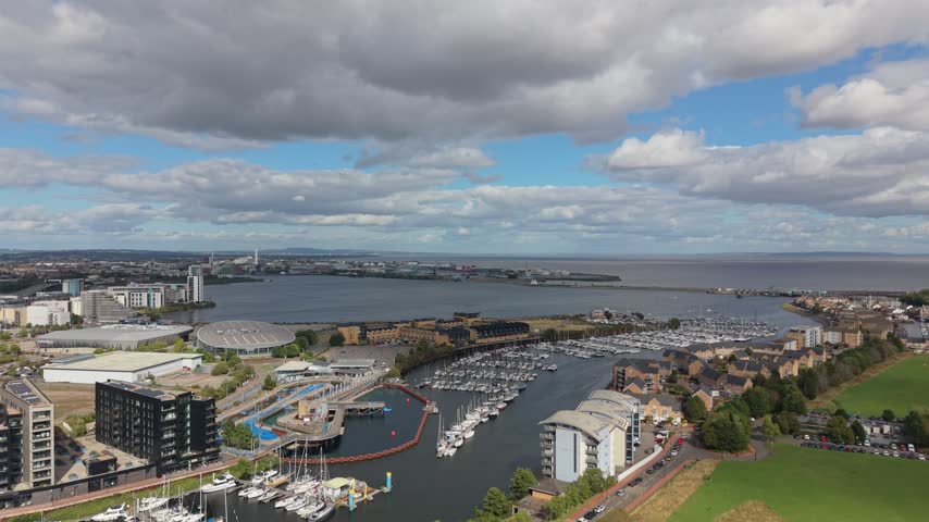 Aerial drone view over Cardiff Bay in Wales, showing a large marina filled with moored yachts and boats, waterfront buildings and the surrounding bay under dramatic clouds.