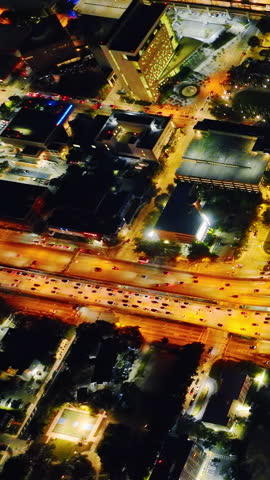 Overhead view of Miami street intersection traffic at night, Florida. Illuminated cars stream along highway beside dark buildings and trees. Vertical video.