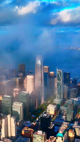 Cloud covered skyline with a tall glass tower, San Francisco. Low fog drifts over downtown high rises beside the bay under a dramatic blue sky. Vertical video.
