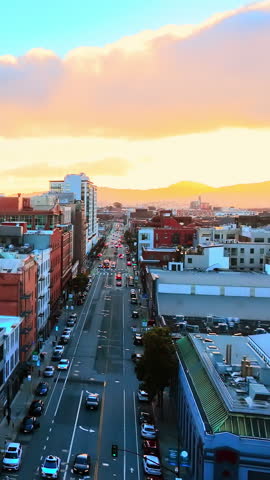 Aerial view of a San Francisco street at sunset, California. Overhead drone shot shows traffic, rooftops and hills glowing under golden evening light. Vertical video.