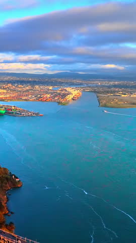 Aerial view of Oakland port and container terminal, California. Overhead drone shot of cargo crane, teal water, coastal cliff and distant hills at dusk. Vertical video.