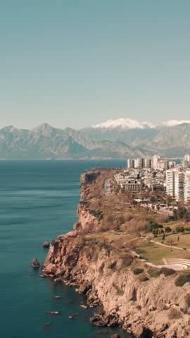 An aerial view displays the coastal city of Antalya built atop rocky cliffs along the Mediterranean Sea. Snow-capped mountains rise in the distant horizon under a clear sky.
