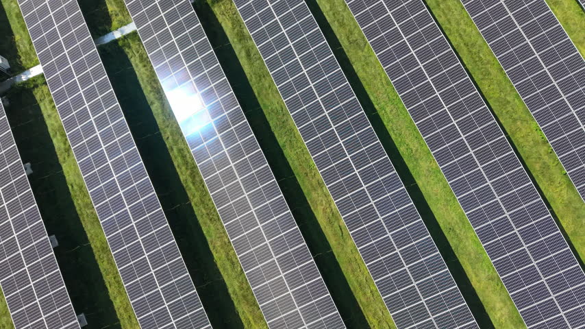 Dense rows of solar panels stretch across flat open terrain, wildflower strips separate angled panel banks, closely spaced grid pattern spans large solar farm in USA. Aerial drone top down