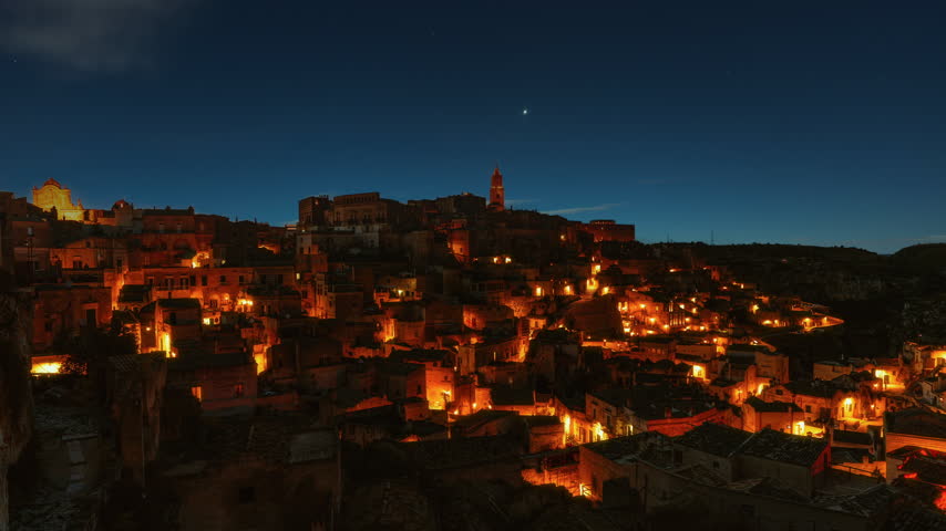 Video loop time lapse of clouds and stars moving over illuminated ancient old city of Matera at night.