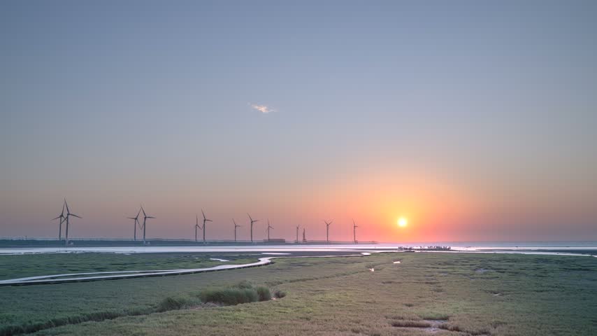 Scenic sunset view over Gaomei Wetland in Taichung, Taiwan, featuring silhouettes of wind turbines and wetland grass landscape.