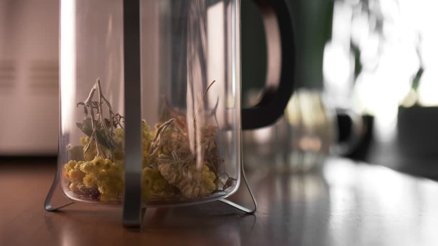 Close-up shot of dried yellow flowers and organic herbs inside a glass french press teapot placed on a wooden table. Warm natural sunlight creating rich contrast and soft blurred background with dried herbal bunches.