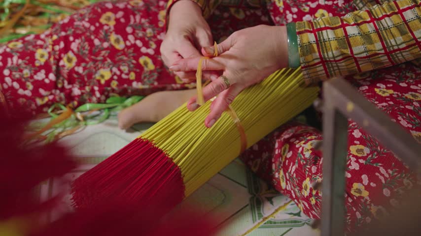 Worker gathers and ties finished yellow and red incense sticks into a compact bundle after drying. Close-up view shows the manual bundling stage used to organize incense before final packaging.