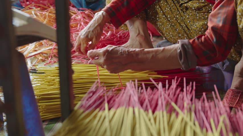 Worker gathers and ties finished yellow and red incense sticks into a compact bundle after drying. Close-up view shows the manual bundling stage used to organize incense before final packaging.