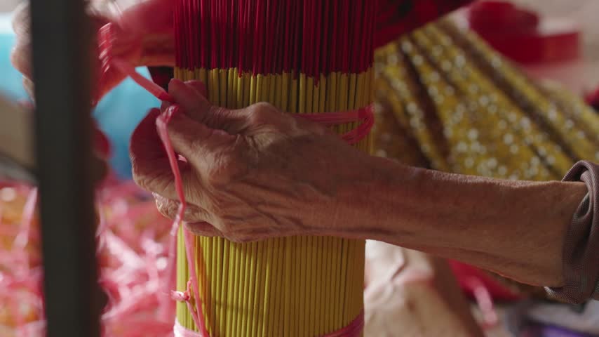 Worker gathers and ties finished yellow and red incense sticks into a compact bundle after drying. Close-up view shows the manual bundling stage used to organize incense before final packaging.