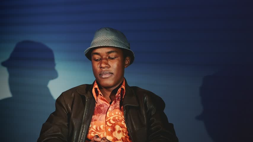 An African-descent man wearing a leather jacket and fedora speaks and gestures indoors with a dark blue-gray gradient background and a faint silhouette behind him.