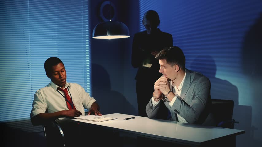 Two men sit at a table discussing papers while a woman stands behind them observing in an office room with window blinds and dramatic lighting.