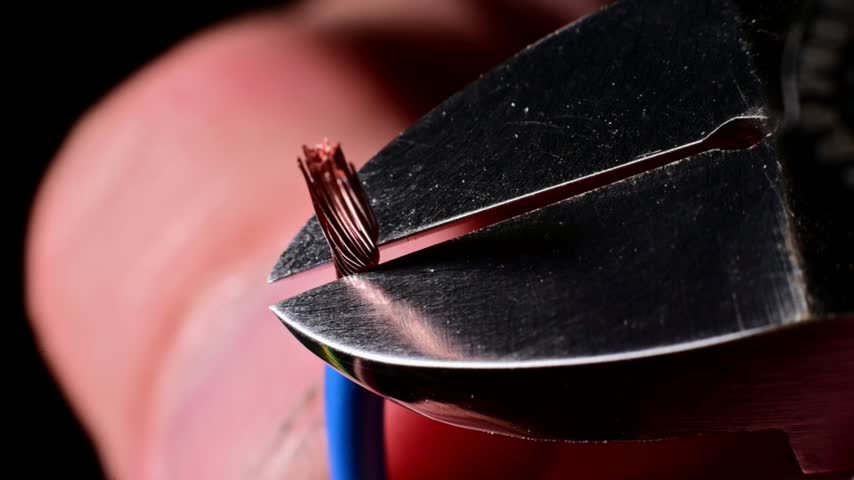 Metal wire cutters sever stranded copper wire in a close-up slow-motion shot. The dark indoor setting features shallow depth of field with metallic debris.
