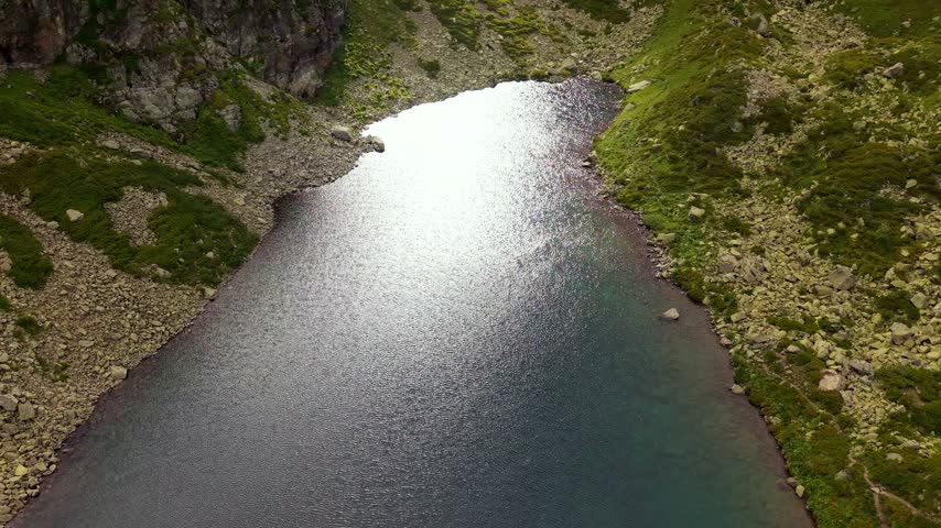 An aerial view of a mountain lake surrounded by rocky slopes. The camera ascends and tilts down, revealing the water surface and terrain. Natural light reflects off the water.