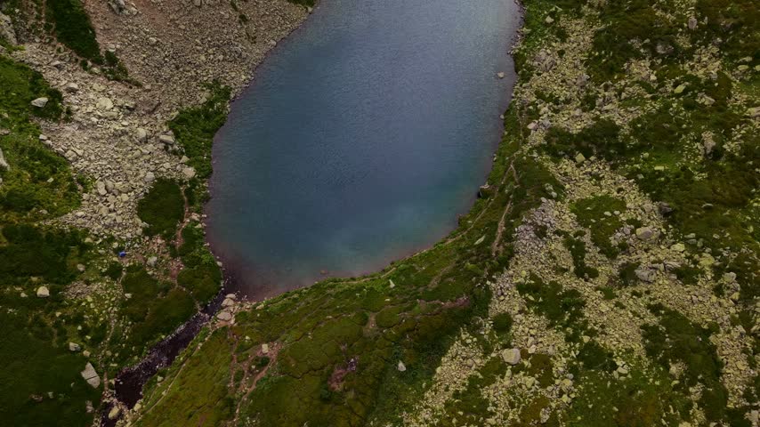 An aerial view pulls backward from a mountain lake. The rocky alpine environment features green vegetation and a small tent near the shore. Natural daylight illuminates the landscape.