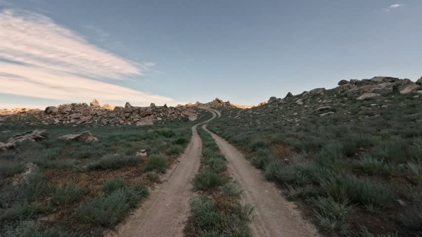 Forward motion along a dirt road winding through a grassy, rocky landscape. The camera moves steadily toward a small ridge under a clear sky. Natural daylight illuminates the terrain.