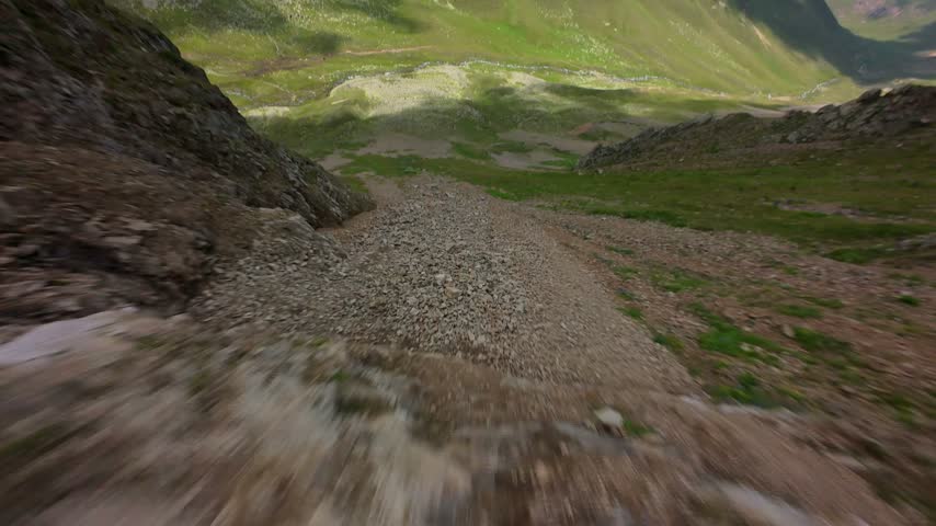 Fast forward perspective descending a steep mountain slope. The view moves from rocky scree to grassy terrain with small flowers. Natural daylight illuminates the landscape.