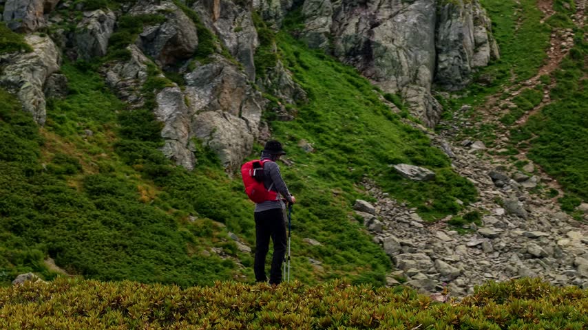 A woman in hiking gear stands on a mountain ridge and looks upward. The medium shot features natural daylight in a rocky environment. The static camera captures the scene in slow motion.