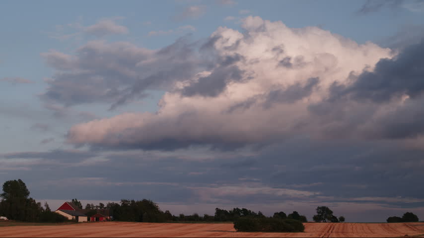 A massive cumulus cloud builds over quiet farmland with a red barn and scattered trees at dusk. Shifting light and shadow move across the fields beneath the developing storm.