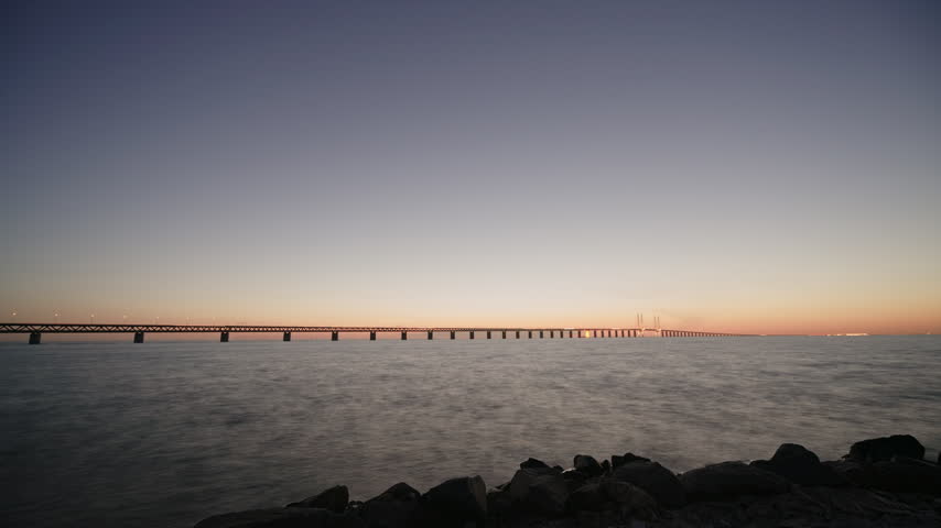 The illuminated Öresund Bridge spans the strait at twilight. An aircraft leaves a red and green light trail across the dark sky above the glowing horizon.