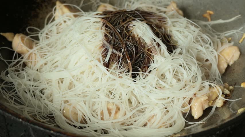 A slow-motion close-up video capturing a spatula stir-frying seasoned rice vermicelli noodles with large chunks of tender chicken meat in a hot wok, showing slight steam and oily texture details during Asian-style home cooking.