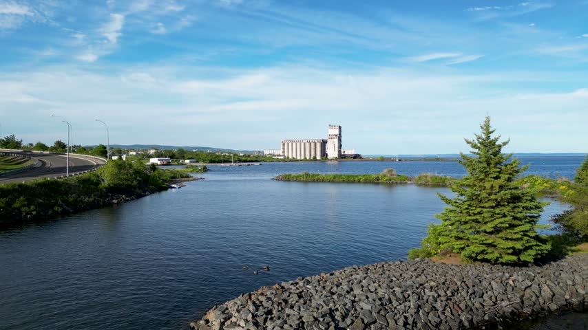 Drone flies forward over calm blue lake water toward an industrial grain elevator along the Thunder Bay waterfront in Ontario, Canada.