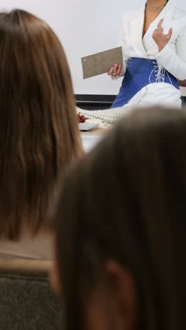 Female presenter speaking to audience in classroom, holding clipboard and gesturing during seated lecture, students seen from behind, clean white wall background behind speaker and audience.