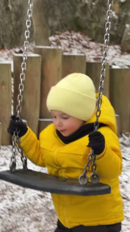 A smiling toddler boy holds the chains of a swing and plays beside its seat with his mother nearby in snowy Bednarski Park, Krakow.