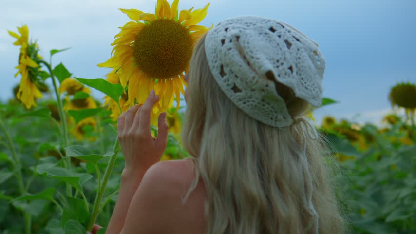 Girl with long blonde hair and a white crocheted hat gently touches a sunflower. She stands in a vast sunflower field under soft sky. Sunflowers bloom brightly, their yellow petals facing the sun