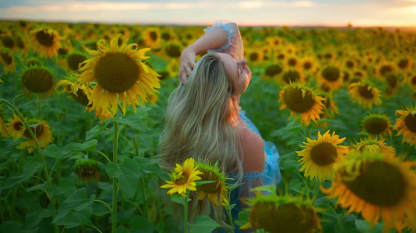 Woman in blue dress stands amid tall sunflowers. Sunlight glows warmly around her. She closes eyes, enjoying peaceful moment. Field stretches endlessly to horizon. Perfect for travel, wellness