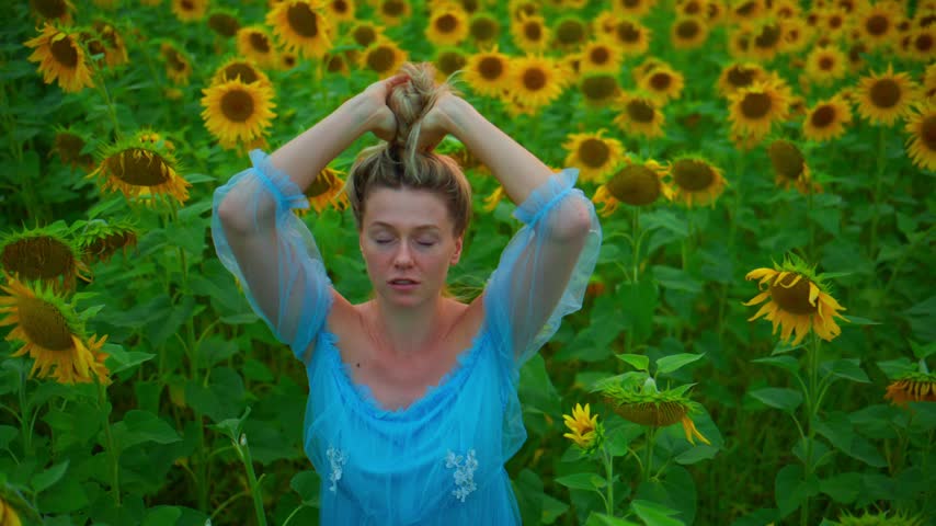 Woman in blue dress stands among sunflowers. She raises arms, hair tied, eyes closed. Feels peaceful, connects with nature. Ideal for travel, wellness, or summer-themed projects