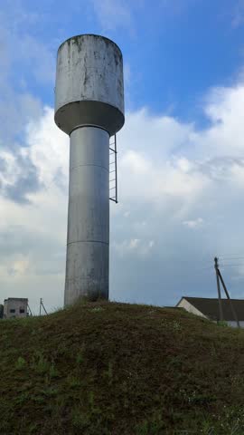 Tall water tower with a cylindrical body and round top, positioned on a grassy hill, with distant structures and power lines visible in the background