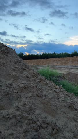 Excavation site features a prominent dirt mound and a green truck on sandy ground, with a backdrop of trees and cloudy sky, illustrating construction activities
