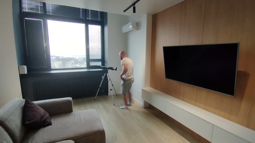A man looks through a telescope mounted at a large window in a modern apartment, admiring the cityscape.