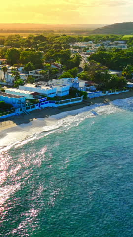 White beachfront houses along a sandy shore, Puerto Rico. Elevated view at sunset with golden sky, sparkling teal water, foamy waves, palm trees and green hills in the distance. Vertical video.
