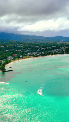 Jet skis on turquoise bay under stormy clouds, Puerto Rico. Elevated view of three watercraft leaving white wakes, curved sandy shoreline, town and green mountains beneath gray sky. Vertical video.