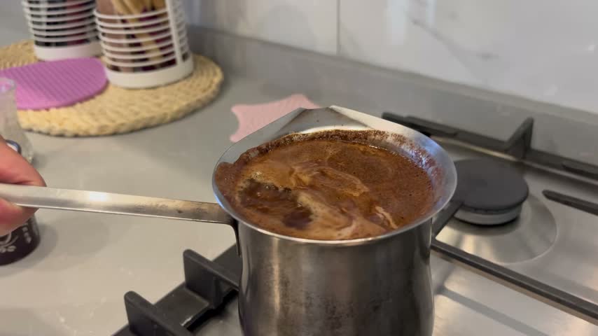 Close-up of a person holding the handle of a traditional cezve pot filled with foaming hot Arabic coffee over a stove.