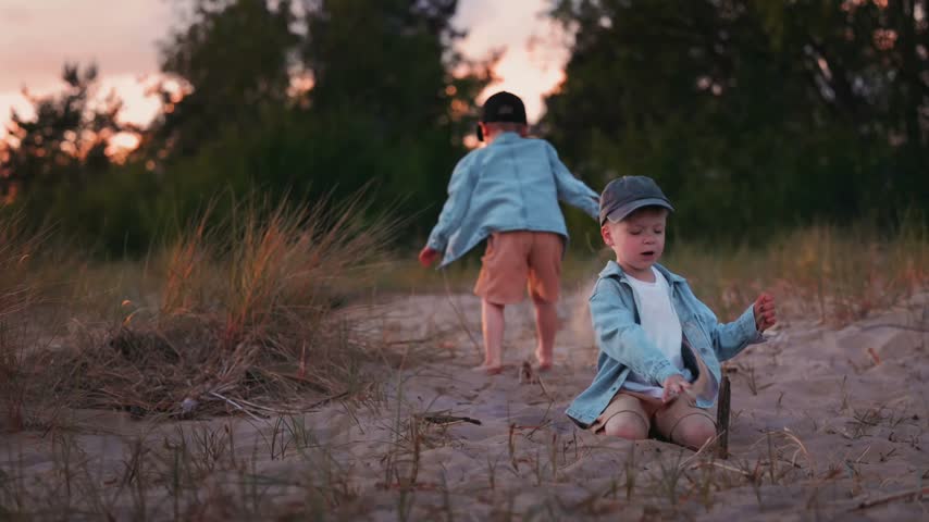 Two boys interact at the river shore, one seated on the sand and the other standing, both dressed in light summer clothing amidst grass and trees