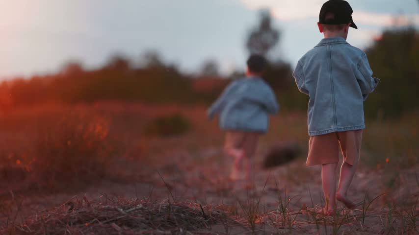 Two boys at a river shore, one in a denim jacket and cap, walking through sandy terrain with grass and trees in the background