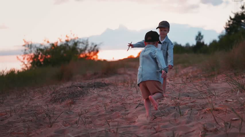 Two boys in denim jackets interact playfully by the river shore during sunset, surrounded by grass and trees, capturing a moment of childhood joy