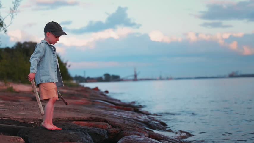 Young boy stands on river shore, barefoot on rocks, holding a stick while looking at the water, with trees and boats in the background