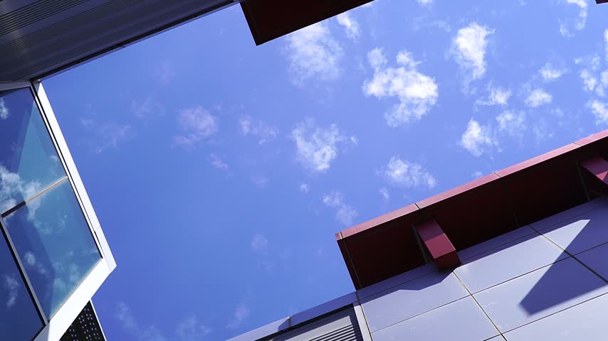 Clouds drift across a clear blue sky, viewed from below and framed by the sharp angles of contemporary office buildings