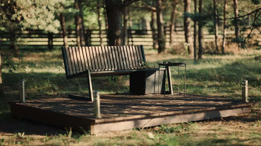 A bench and a table are placed on a wooden platform in a forest. The sun shines through the trees. Grass grows around the area with some scattered plants.
