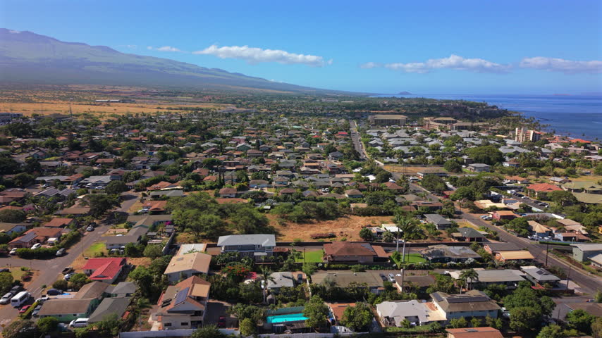 Aerial view of Kihei on Maui in the Hawaii island chain showing tropical suburban homes palm trees and green lawns with the West Maui mountains and blue Pacific coastline behind.