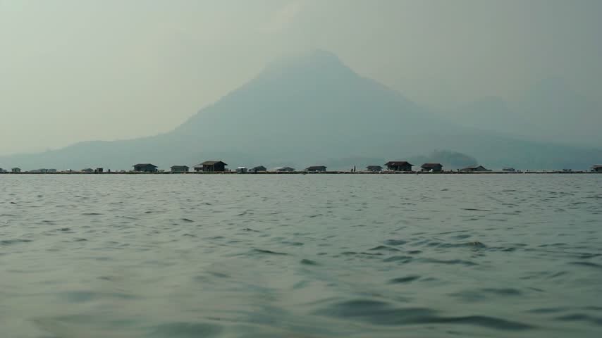 A wide shot of gentle water ripples on a large lake surface, featuring traditional floating fish farm cages, wooden huts, and a misty mountain silhouette under a hazy sky.